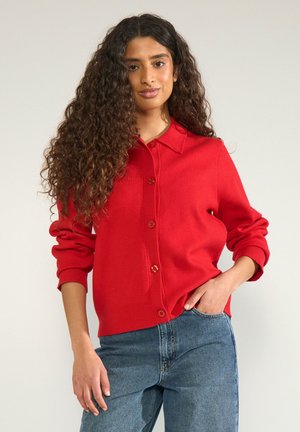 Femme aux longs cheveux bouclés portant un cardigan rouge boutonné et un jean bleu, debout avec une main dans la poche, devant un fond uni.