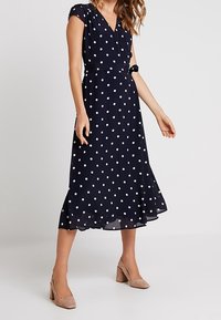 Granatowa sukienka midi w groszki z krótkimi rękawami, kopertowym gorsem i rozkloszowanym dołem. Zapinana na wiązanie w talii, wykonana z lekkiego materiału.