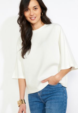 Žena s dlhými tmavými vlasmi, oblečená v voľnej bielej blúzke a modrých džínsoch, usmievajúca sa, s jednou rukou vrecku a zlatými kruhovými náušnicami a náramkom.