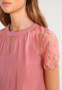 Närbild av en kvinna som bär en topp i rosa med spetsärmar, med en nyckelhålsutskärning, som framhäver en mjuk och feminin stil.