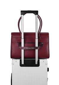 Un sac à main texturé rouge repose sur une valise rigide blanche, maintenue par une poignée en aluminium rétractable, arborant une étiquette de marque.