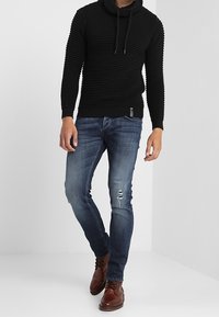 Man in een gestructureerde zwarte hoodie, slanke blauwe spijkerbroek met een verstoord gedeelte, en bruine leren veterschoenen, staande tegen een witte achtergrond.