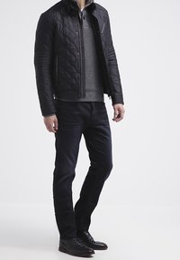 Uomo che indossa una giacca trapuntata nera sopra un maglione di lana grigia, pantaloni scuri e scarpe eleganti nere, in piedi contro uno sfondo bianco.