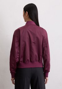 Woman with long dark hair wearing a maroon bomber jacket and black pants, facing a plain white wall, back shown.