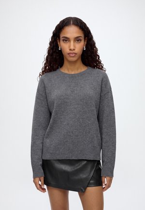 Femme aux longs cheveux bouclés portant un pull gris et une mini-jupe en cuir noire, debout devant un fond blanc uni.