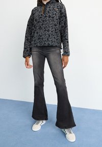 Fleece pullover in dark gray with a textured leaf pattern, half-zip collar, and brown accents. Paired with faded black flared jeans and sneakers.