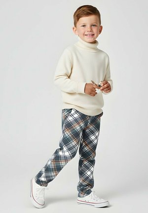 Młody chłopiec uśmiechający się, ubrany w kremowy golf, kratkowane spodnie i białe tenisówki, trzymający małą figurkę zabawki z skrzydłami.