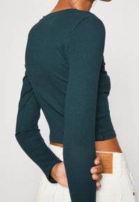 Tiefgrünes langärmliges Rippenoberteil mit anliegendem Design, Rundhalsausschnitt und cropped Länge, kombiniert mit hellen Jeans, die ein Etikett präsentieren.