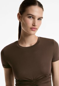 T-shirt marrone attillato con maniche corte, scollo rotondo, dettagli arricciati in vita, realizzato in tessuto morbido e elasticizzato.