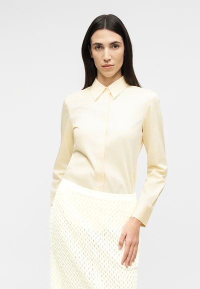 Γυναίκα με μακριά σκούρα μαλλιά, ντυμένη με κρεμ πουκάμισο με κουμπιά και υφασμάτινη φούστα σε κρεμ απόχρωση, στέκεται μπροστά σε απλό ανοιχτό φόντο.