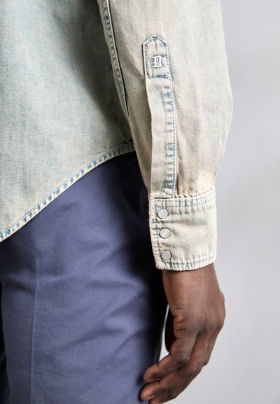 Camisa de mezclilla azul claro con dobladillo curvado, que presenta tres cierres de botones en el puño y bordes deshilachados en la manga. Pantalones azul oscuro.