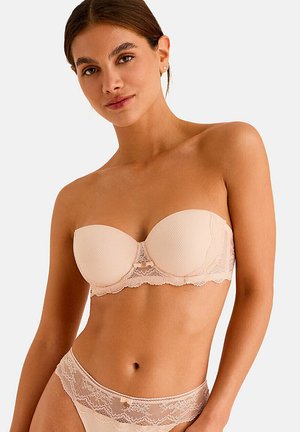 Jeune femme portant un soutien-gorge bustier en dentelle beige et une culotte assortie en dentelle, posant devant un fond clair uni.