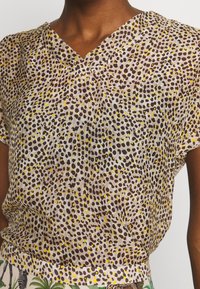 Bluzka z krótkim rękawem z przezroczystego materiału, z dekoltem w kształcie V, w żółte, brązowe i czarne kropki na jasnym tle.