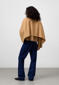 Beige wolmix cape met een ronde zoom, gecombineerd met donkerblauwe wijde jeans. De outfit heeft een ontspannen, gelaagd silhouet.