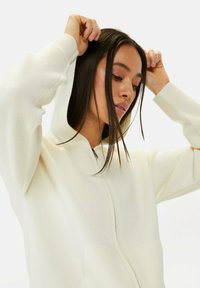 Witte zip-up hoodie van een gestructureerde stof, met een capuchon en geribde manchetten, gedragen door een persoon met lang haar.