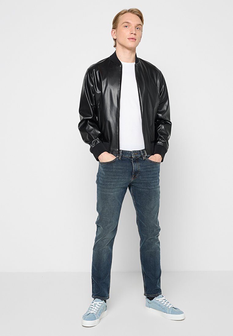 Jeune homme debout, les mains dans les poches, portant une veste en cuir noire, une chemise blanche, un jean bleu et des sneakers bleu clair sur fond uni.