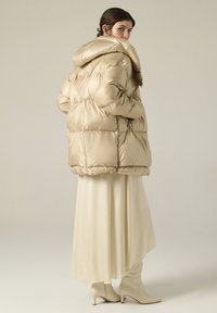 Giacca puffer beige con cappuccio, caratterizzata da un design trapuntato e una texture liscia. Indossata sopra un abito lungo color crema e stivali alla caviglia coordinati.