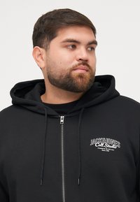 Crna zip-up dukserica s kenguruvim džepom, užadima i izvezenim logotipom na lijevoj strani prsnog područja. Mekani materijal, opušten kroj, minimalistički dizajn.