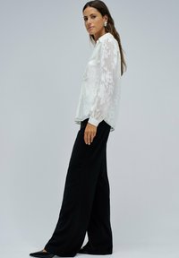 Blusa bianca trasparente con ricami floreali, maniche lunghe e colletto, abbinata a pantaloni neri a vita alta e larghi e scarpe nere a punta.