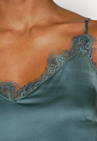 Close-up of a person wearing a teal satin camisole with delicate lace trim along the neckline and spaghetti straps.