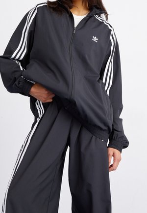 Personne portant un survêtement Adidas noir avec des bandes blanches sur les manches et le pantalon, remontant partiellement la veste.