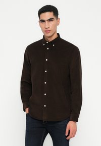 Brun kordfløyelskjorte med krage som kan kneppes, med hvite knapper langs fronten. Langermet og design med buet kant.