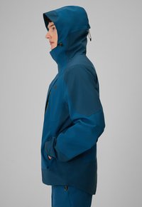 Personne portant une veste bleue à capuche, les mains dans les poches, debout de profil sur un fond gris uni.
