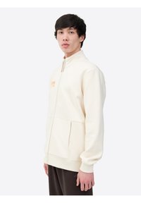 Chaqueta cremosa clara con cierre de cremallera, cuello alto, bolsillos laterales y logo naranja en el pecho. Textura suave y corte casual.