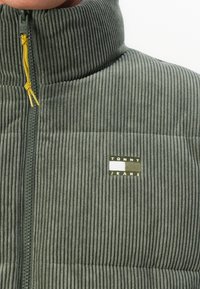 Close-up of ribbed olive green zip-up jacket with high collar, yellow zipper pull, and Tommy Jeans logo on chest.