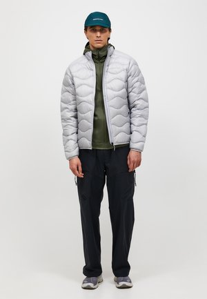 Man draagt een lichtgrijs doorgestikt jack, daaronder een olijfgroen jack met rits, zwarte cargobroek, grijze sneakers en een donkerteal pet, staand tegen een witte achtergrond.