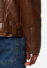 Brun läderjacka med dragkedja vid ärmslut, tryckknappsstängning och kviltad textur, i kombination med blå denimjeans.
