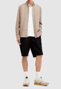 Camisa de botón color marrón claro, camiseta de tirantes blanca de canalé, pantalones cortos negros, calcetas blancas y zapatillas beige con acentos texturizados.