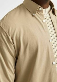 Beige skjorta med knappar och en texturerad krage, lager-på-lager över en vit stickad tröja med rutmönster. Närbild av tyg och detaljer.