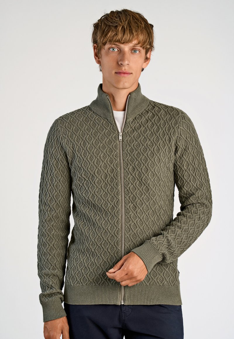 Kaki grön zip-up tröja med texturerat diamantmönster, ribbad krage och muddar, samt en slät dragkedja. Bärs över en vit skjorta.