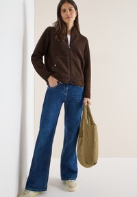 Jeune femme portant une veste polaire marron, un jean large bleu, des baskets blanches, et tenant un sac fourre-tout matelassé beige.