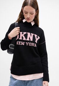 Czarny sweter z dzianiny z różowym napisem "DKNY NEW YORK", prążkowanymi mankietami i dekoltem. Noszony na jasnoróżowej koszuli z kołnierzykiem. Widoczne dżinsowe spodnie.