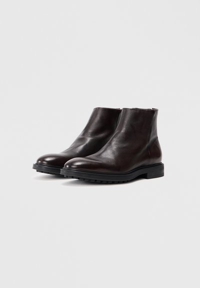 Par de botas de tornozelo de couro castanho escuro com solas de borracha pretas sobre um fundo cinza claro liso.