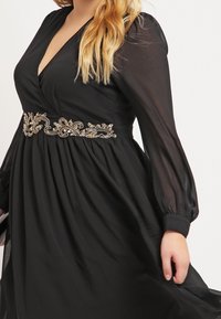 Czarna sukienka o kroju owijanym z długimi przezroczystymi rękawami, głębokim dekoltem w serek i ozdobnym paskiem z koralikami w kwiatowy wzór podkreślającym talię. Zwiewny materiał o teksturowanym wykończeniu.