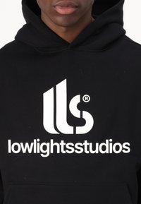 Felpa nera con cappuccio, caratterizzata da un prominente logo grafico bianco con il testo "lowlightstudios" e forme astratte, tessuto texturizzato e design con cappuccio.