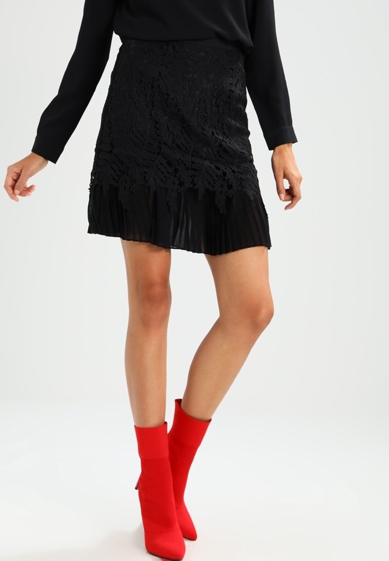 Femme portant une jupe en dentelle noire avec un ourlet plissé et des bottines rouges vives, debout devant un fond clair uni.