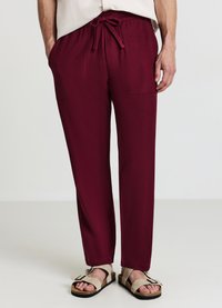 Pantaloni bordeaux a vestibilità rilassata con vita a coulisse, due tasche frontali e una texture liscia, abbinati a sandali beige.