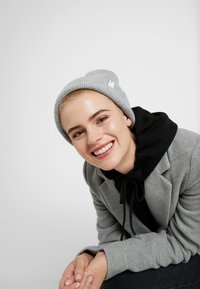 Gray knit beanie with a small tag, worn with a black hooded sweatshirt and a gray blazer. Smooth texture, simple design, and snug fit.