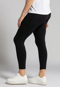Leggings noirs fabriqués à partir d'un tissu extensible et léger. Présentent un design ajusté avec une texture lisse et une coupe à la cheville, associés à des baskets blanches.
