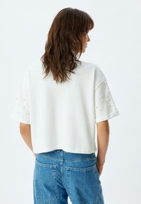 Witte korte mouwen top met getextureerde parelaccenten op de mouwen en een cropped zoom. Draagt met blauwe high-waisted jeans.