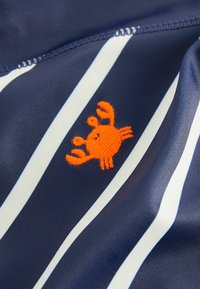 Tissu bleu marine avec des rayures verticales blanches ; présente un design de crabe brodé orange près de l'ourlet. Le matériau semble lisse et léger.