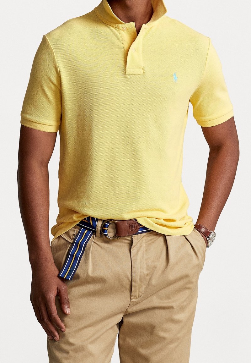 Homme portant un polo jaune à manches courtes rentré dans un pantalon beige avec une ceinture rayée bleue et une montre marron au poignet gauche.