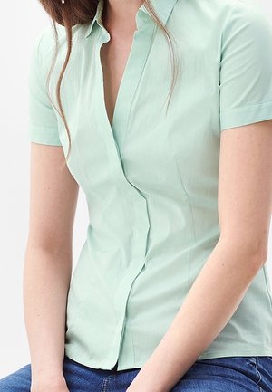 Mujer con una blusa ajustada de manga corta color verde claro y jeans azules, sentada con los brazos descansando en su regazo.