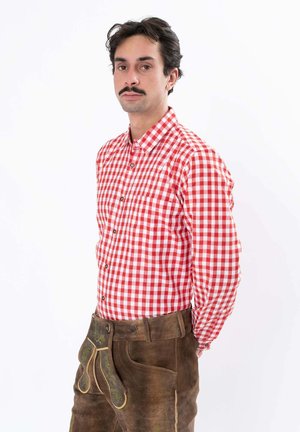 Mann mit Schnurrbart trägt rot-weiß kariertes Hemd und traditionelle braune Lederhose mit bestickter Klappe, steht vor weißem Hintergrund.