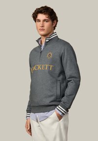 Grijze fleece pullover met kwart-rits, gouden "HCKETT" tekst en geborduurd logo. Heeft ribgebreide boorden met zwart-witte strepen.