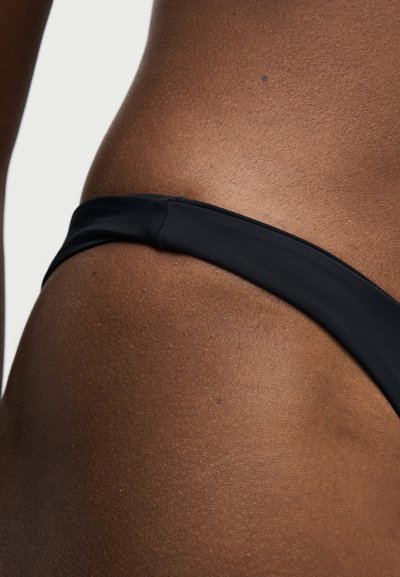 Κοντινό σεγόνου και άνω μηρού ενός ατόμου που φορά μαύρο κάτω μέρος μαγιό σε απλό φωτεινό φόντο.